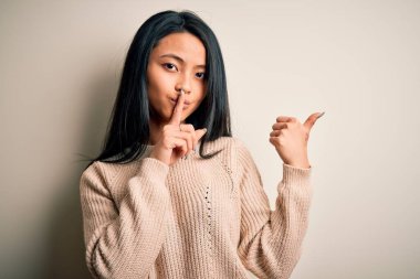 İzole edilmiş beyaz arka plan üzerine gündelik süveter giyen genç, güzel Çinli bir kadın ellerini yana uzatarak parmaklarını dudaklarına uzatarak sessiz olmak istiyor. Sessizlik ve gizli konsept.