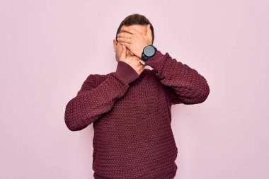 Mavi gözlü genç adam pembe arka planda dikilmiş süveter giyiyor gözleri ve ağzı kapalı, şaşırmış ve şok olmuş. Duygularını gizliyor.