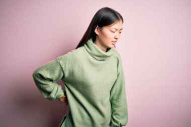 Genç, güzel Asyalı bir kadın pembe arka planda yeşil bir kazak giyiyor sırt ağrısı çekiyor, elleriyle dokunuyor, kas ağrısı çekiyor.