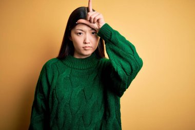 Sarı arka plan üzerine yeşil kazak giyen genç, güzel Asyalı bir kadın. Alnında parmakları olan insanlarla alay ediyor. Ezik hareketler yapıyor, alay ediyor..