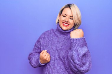 Güzel sarışın ve cüsseli bir kadın mor arka plan üzerine balıkçı yaka kazak giyip kolları havada ve gözleri kapalı bir şekilde başarıyı kutluyor. Kazanan konsept.