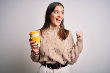 Genç, güzel esmer kadın izole edilmiş beyaz arka planda bir bardak kahve içiyor ve gülümseyen yüzüyle baş parmağıyla işaret ediyor.