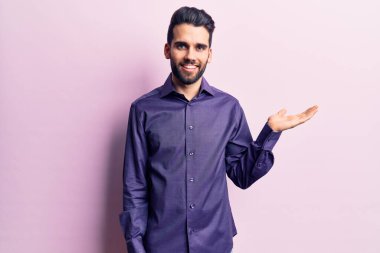 Sakallı, sıradan tişörtlü, neşeli gülümseyen, eli avucunda kameraya bakan genç ve yakışıklı bir adam.. 