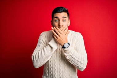 İzole edilmiş kırmızı arka planda duran beyaz süveter giyen genç yakışıklı adam yanlışlıkla ağızlarını kapatarak şok etti. Gizli konsept.