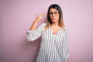 Genç ve güzel bir kadın, pembe arka planda günlük çizgili tişört ve gözlük takıyor. Kendini vuruyor ve öldürüyor. Silah gibi el ve parmaklarını işaret ediyor..