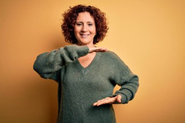 Orta yaşlı, kıvırcık saçlı güzel bir kadın izole edilmiş sarı arka plan üzerine gündelik süveter giyiyor. Elleri büyük ve büyük boy işareti, ölçü sembolü gösteriyor. Kameraya bakıp gülümsüyor. Ölçüm konsepti.