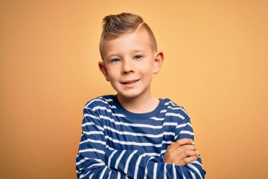 Mavi gözlü, sarı arka plan üzerinde deniz çizgili tişört giyen, kollarını kavuşturup kameraya bakan, gülümseyen küçük beyaz bir çocuk. Pozitif kişi.