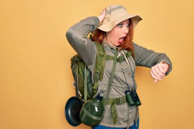 Sırt çantalı, sarı şapkalı, sırt çantalı, genç, kızıl saçlı bir kadın geç kalmaktan korkarak saatine bakıyor.