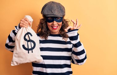 Orta yaşlı esmer soyguncu kadın maske takıyor ve elinde dolar sembolü olan bir çanta tutuyor. Parmakla işaret ediyor, gülümsüyor ve mükemmel bir sembol oluşturuyor.