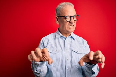 Orta yaşlı yakışıklı beyaz bir adam kırmızı arka planda günlük çizgili gömlek ve gözlük takıyor komik bir şekilde kedi gibi pençe hareketi yapıyor, agresif ve seksi bir ifade.