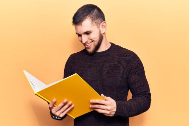 Kitap okuyan yakışıklı genç adam mutlu ve pozitif görünüyor. Kendine güvenen bir gülümsemeyle ayakta duruyor ve dişlerini gösteriyor. 