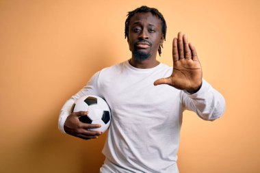 Genç Afrikalı Amerikalı oyuncu futbol topunu sarı arka planda tutarak oynuyor. Açık elle dur işareti yapıyor. Ciddi ve kendinden emin bir ifadeyle, savunma hareketi yapıyor.