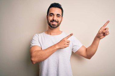 Sakallı, sıradan tişörtlü genç bir adam beyaz arka planda dikiliyor gülümsüyor ve kameraya bakıyor iki eliyle ve parmaklarıyla yan tarafa bakıyor..