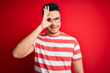 Sıradan çizgili tişört ve gözlük takan genç yakışıklı adam izole edilmiş kırmızı arka planda el hareketleriyle gülümsüyor ve mutlu yüzle parmaklarının arasından bakıyor..