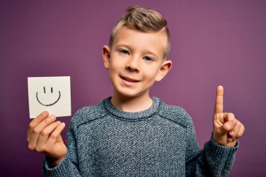 Bir kağıda gülen surat çizen küçük beyaz çocuk bir fikir ya da soruyla şaşırmış gibi mutlu bir mesaj veriyor.