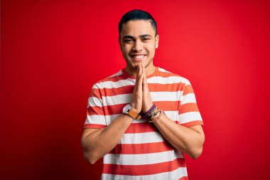 Sıradan çizgili tişört giyen genç Brezilyalı bir adam izole edilmiş kırmızı arka planda duruyor ellerini birleştirip af diliyor kendinden emin bir şekilde gülümsüyor..