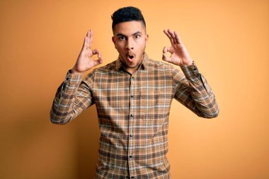 İzole edilmiş sarı arka plan üzerinde duran sıradan tişört giyen genç ve yakışıklı bir adam parmaklarıyla onay sembolü yaparken şaşırmış ve şok olmuş görünüyor. Çılgın ifade