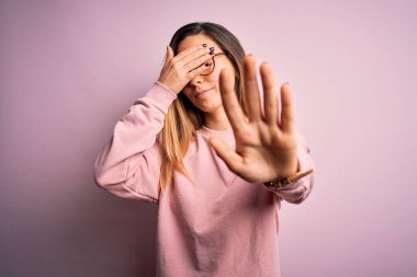 Mavi gözlü güzel sarışın kadın pembe arka plan üzerine süveter ve gözlük takıyor gözleri ellerle örtüyor ve üzgün ve korku ifadeleriyle dur hareketi yapıyor. Utanç verici ve olumsuz bir kavram..