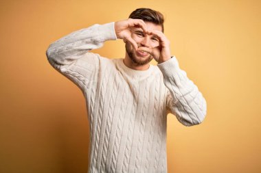 Sarışın, sakallı, mavi gözlü, sarı arka planda beyaz süveter giyen el ve parmakları ile kalp şeklinde gülümseyen genç bir adam.