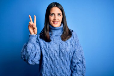 Mavi gözlü, balıkçı yaka kazak giyen genç esmer kadın kameraya bakıp zafer işareti yaparken gülümsüyor. İki numara..