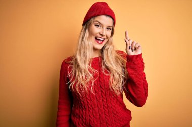 Genç, güzel, sarışın, sıradan bir süveter giyen, beyaz arka planda yün şapkalı, başarılı bir fikri işaret eden bir kadın. Heyecanlı ve mutlu. Bir numara..