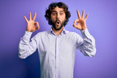 Mor arka plan üzerinde duran sakallı yakışıklı iş adamı şaşırmış ve şaşırmış görünüyor. Tamam sembolünü parmaklarıyla yapıyor. Çılgın ifade