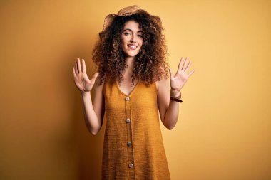 Güzel esmer kadın tatilde. Kıvırcık saçlı, piercing takan, şapka takan, elbise giyen, on numaralı parmaklarıyla işaret eden, kendinden emin ve mutlu bir şekilde gülümseyen..