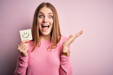 Genç, güzel, kızıl saçlı kadın elinde gülümseyen emoji mesajıyla mutlu ve heyecanlı, kazanan yüz ifadesi zaferi büyük bir gülümsemeyle kutluyor ve ellerini kaldırıyor.