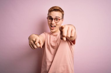 Genç ve yakışıklı kızıl tişörtlü adam izole edilmiş pembe arka planda dikiliyor. Seni ve kamerayı parmaklarıyla gösteriyor, neşeli ve pozitif gülümsüyor.