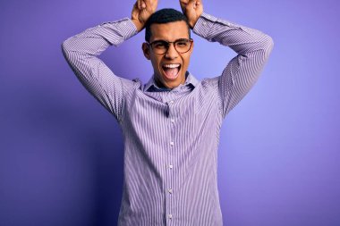 Yakışıklı Afro-Amerikan adam çizgili gömlek ve gözlük takıyor mor arka planda komik ve çılgın pozlar veriyor parmakları kafasında tavşan kulağı gibi, neşeli gülüyor.