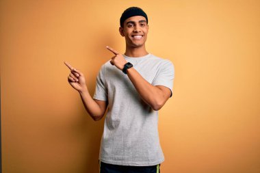 Yakışıklı Afro-Amerikan sporcu sarı arka planda spor giysisi giyiyor gülümsüyor ve kameraya bakıyor iki eliyle ve parmaklarıyla yandan işaret ediyor..