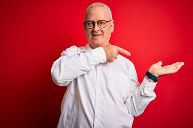 Orta yaşlı, beyaz gömlekli, kırmızı arka planda gözlüklü yakışıklı adam kameraya hayret ediyor ve gülümsüyor..