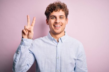 Sarışın, kıvırcık saçlı, beyaz arka planda çizgili tişörtlü, genç, yakışıklı bir adam. İki numaralı parmaklarıyla işaret ediyor. Kendinden emin ve mutlu gülümsüyor..