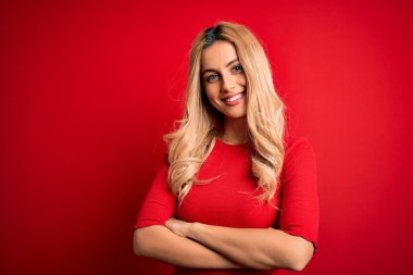 Young beautiful blonde woman wearing casual t-shirt standing over isolated red background happy face smiling with crossed arms looking at the camera. Positive person.