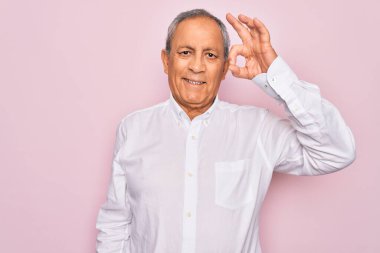 Üst düzey yakışıklı, gri saçlı, zarif tişörtlü, pembe arka planda, gülümseyen, el ve parmaklarıyla tamam işareti yapan bir adam. Başarılı ifade.