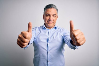 Orta yaşlı, yakışıklı, gri saçlı iş adamı beyaz arka planda zarif gömlekler giyiyor. Elle olumlu jest yapıyor, gülümsüyor ve başarı için seviniyor. Kazanan jest.