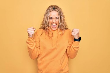 Sarışın, güzel ve sportif bir kadın sarı arka plan üzerine sıradan bir kazak giyiyor kızgın ve kızgın yumruklar savuruyor öfkeyle bağırırken sinirli ve öfkeli. Öfke ve agresif kavram.