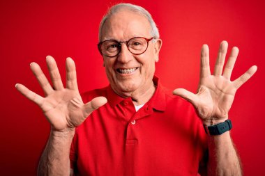 Gri saçlı, gözlüklü, kırmızı arka planda günlük tişörtlü, kendinden emin ve mutlu bir şekilde gülümseyen on numaralı parmaklarıyla işaret eden son sınıf öğrencisi..