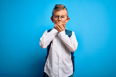 Mavi arka plan üzerinde akıllı gözlük ve okul çantası takan küçük beyaz öğrenci yanlışlıkla ağızlarını kapatarak şok geçirdi. Gizli konsept.