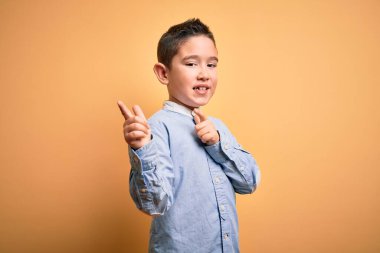 Zarif tişörtlü küçük bir çocuk sarı izole edilmiş arka plan üzerinde durmuş mutlu ve komik yüzlü parmaklarını kameraya doğrultuyor. İyi enerji ve titreşimler.