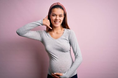 Genç, güzel bir kız hamile, pembe arka planda bebek bekliyor. Gülümsüyor, el ve parmaklarıyla telefonla konuşuyor gibi. İletişim kavramları.