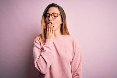 Güzel, mavi gözlü, pembe arka planda süveter ve gözlük takan sarışın kadın ağzıyla ağzını kapatmaktan sıkılmış. Huzursuz ve uykulu..