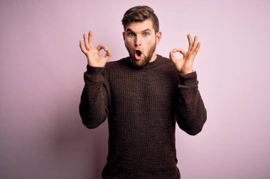 Sakallı ve mavi gözlü genç bir adam pembe arka planda sıradan bir süveter giyiyor. Şaşırmış ve şaşırmış görünüyor. Tamam sembolünü parmaklarıyla yapıyor. Çılgın ifade