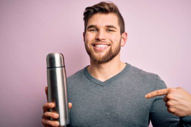 Sarışın, sakallı ve mavi gözlü genç bir adam pembe arka planda termosu olan ve kendini işaret eden sürpriz yüzlü bir adam.