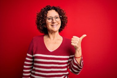 Orta yaşlı, kıvırcık saçlı, sıradan çizgili süveter giyen, kırmızı arka planda gülümseyen, gülümseyen ve baş parmağıyla yüzünü gösteren güzel bir kadın..