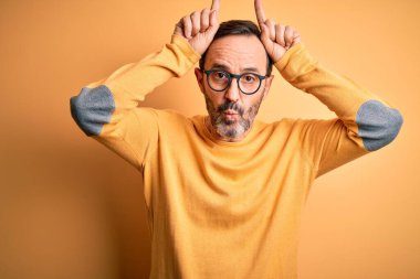Orta yaşlı, ağarmış, günlük süveter ve gözlük takan, izole edilmiş sarı arka plan üzerine komik hareketler yapan, parmağıyla boynuzlarını boynuzlayan bir adam.