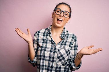 Genç ve güzel esmer bir kadın pembe arka plan üzerine gündelik gömlek ve gözlük takıp çılgınlığı kutluyor ve başarı için hayrete düşüyor. Kolları havada ve gözleri açık, heyecanlı bir şekilde çığlık atıyor. Kazanan konsept