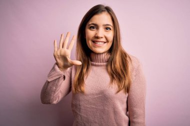Güzel genç bir kadın pembe izole edilmiş arka planda balıkçı yaka kazak giyiyor ve beş numaralı parmaklarıyla işaret ederken kendinden emin ve mutlu bir şekilde gülümsüyor..