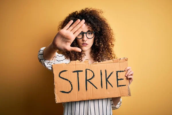 Young beautiful activist woman with curly hair and piercing protesting in strike holding poster with open hand doing stop sign with serious and confident expression, defense gesture