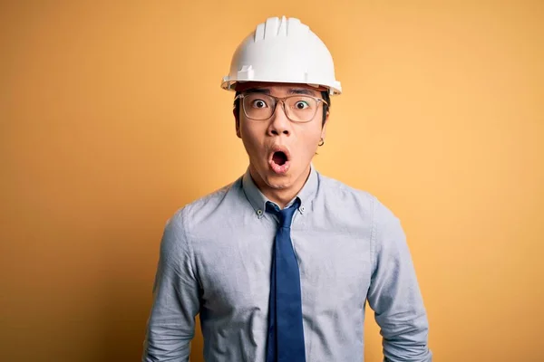 Young handsome chinese architect man wearing safety helmet and tie over yellow background afraid and shocked with surprise expression, fear and excited face.